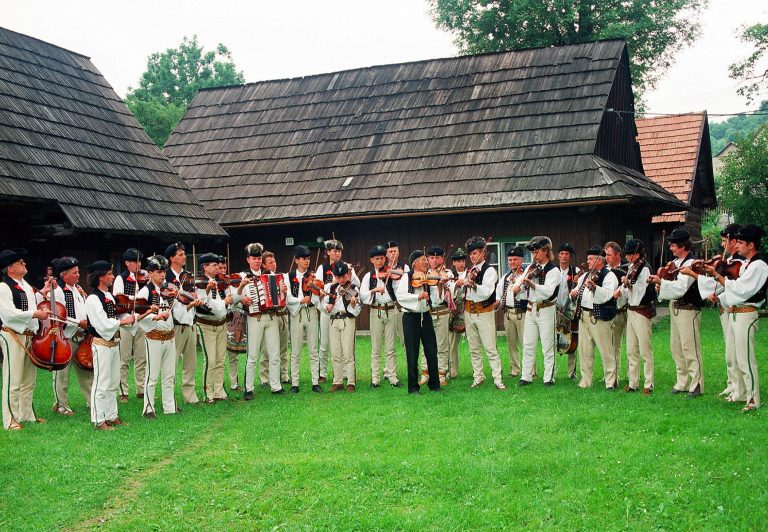 Početná skupina muzikantov v tradičných odevoch hrajúcich na lúke medzi domami.