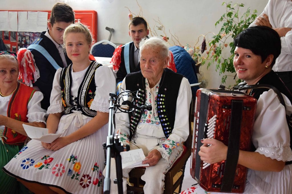Krojovaná akordeónistka  a speváci sedia pred mikfrofónom.