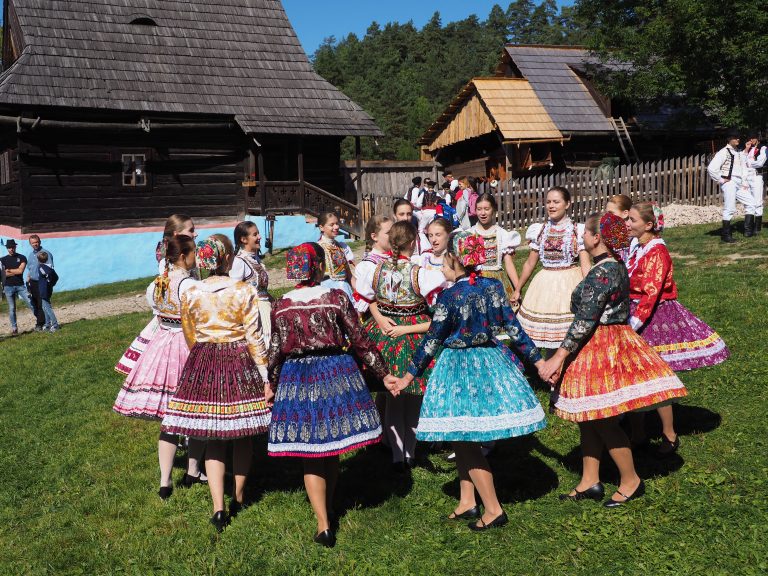 Skupina žien v rôznofarebných tradičných odevoch tancuje kruhový tanec, v pozadí sú drevené stavby.
