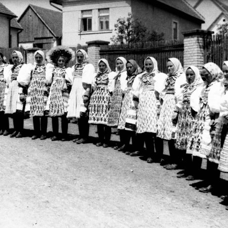 Čiernobiely záber krojovaných žien stojacich na ulici pred domami.