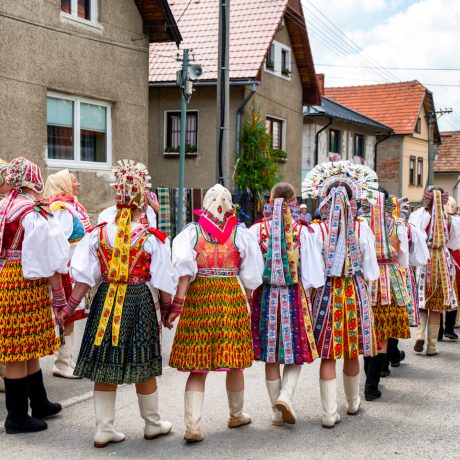 Farebný záber na skupinu krojovaných žien zo zadu na ulici pred domami.