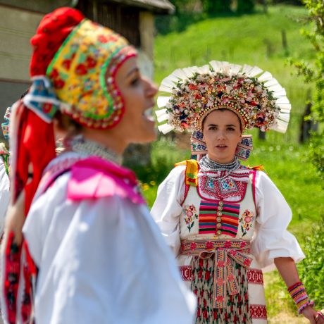 Dve spievajúce krojované ženy, v pozadí zelený trávnatý svah.