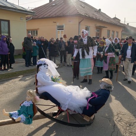Ulica medzi domami plná ľudí, v popredí dve figuríny na kolese.