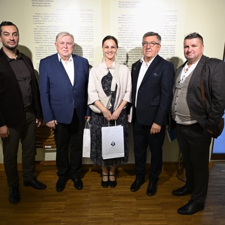Vedľa seba stoja štyria muži a jedna žena, ktorá sa nachádza v strede medzi nimi.