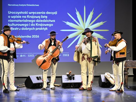 Na pódiu stoja vedľa seba štyria muzikanti v tradičných ľudových odevoch a hrajú na sláčikové nástroje.