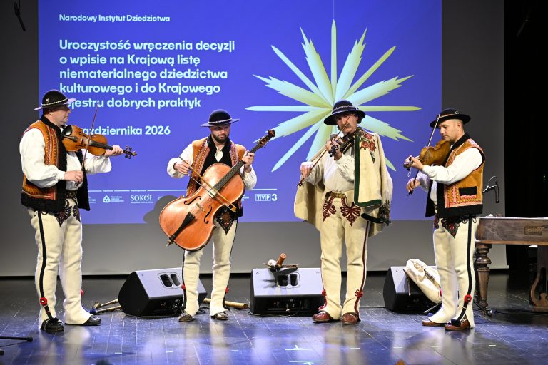 Na pódiu stoja vedľa seba štyria muzikanti v tradičných ľudových odevoch a hrajú na sláčikové nástroje.