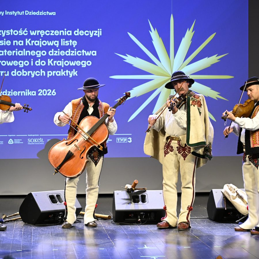 Na pódiu stoja vedľa seba štyria muzikanti v tradičných ľudových odevoch a hrajú na sláčikové nástroje.