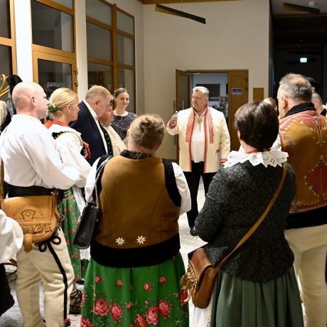 Skupina mužov a žien v tradičných ľudových odevoch stoja v kruhu a venujú pozornosť mužovi v strede.