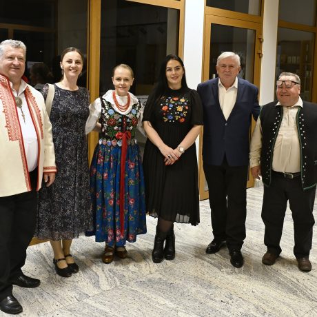 Skupina mužov a žien v tradičnom odeve, ale aj v civilnom spoločenskom odeve stojacich vedľa seba.